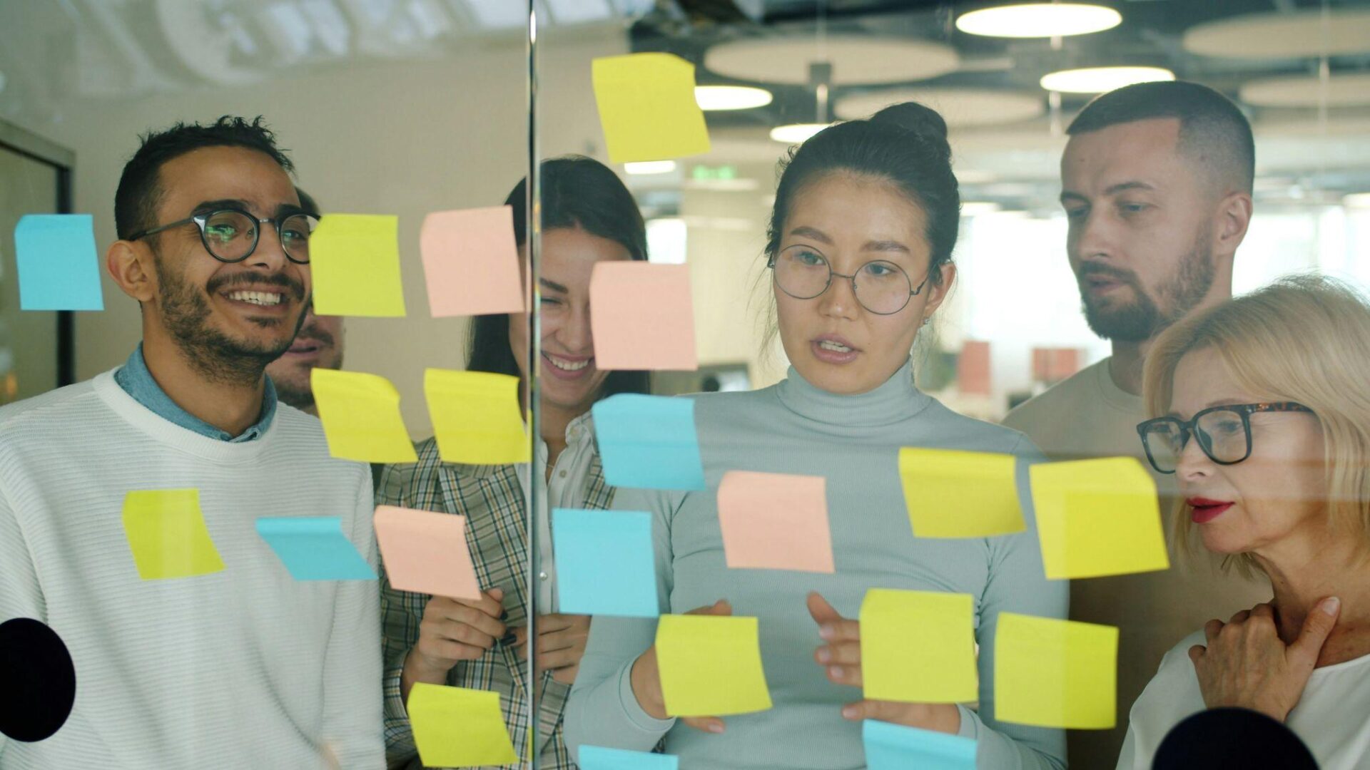 A diverse team of professionals gathered around a glass wall covered with colorful sticky notes, collaborating on ideas and reviewing feedback during a brainstorming session in a modern office